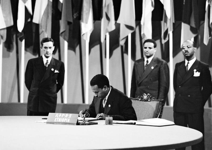 Blata Ephrem Tewelde Medhin Signing the Charter of the United Nations while Vice Minister of Foreign Affairs Aklilu Hapte Wold and Ato Menasse Lema Director General of the Ministry of Finance standing behind him